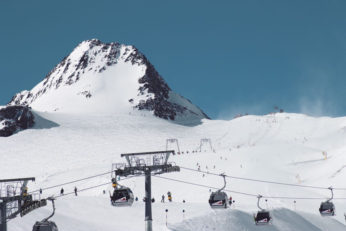 Gondolas rising over a wide, sunlit glacier slope with skiers below a snowy rocky peak