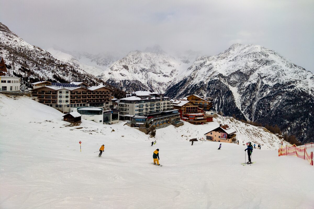The hotels of Hochsölden above a wide groomed slope, with skiers below and snowy peaks behind