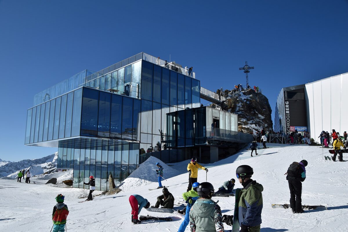 The glass Ice Q restaurant on the Gaislachkogl summit with a cross on the rock behind it, and skiers and snowboarders in the snow in front