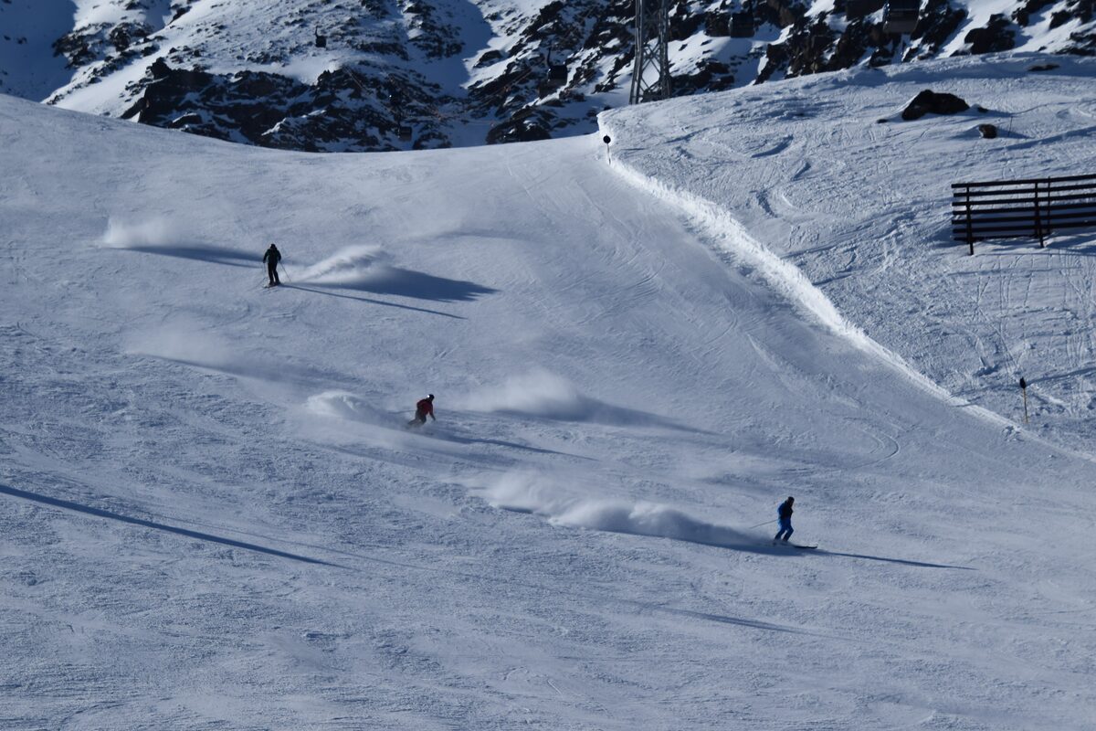 Three skiers carving down a wide groomed slope, with snow spraying off their edges and a lift pylon on the rocky ridge above