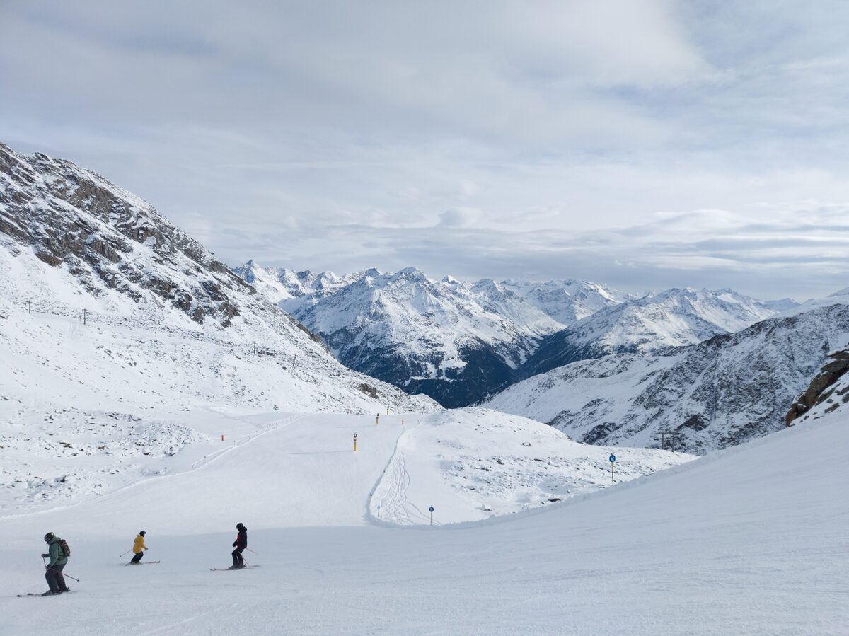 Three skiers on a wide groomed slope below the Schwarzkogl ridge, with a range of snow-covered peaks in the distance
