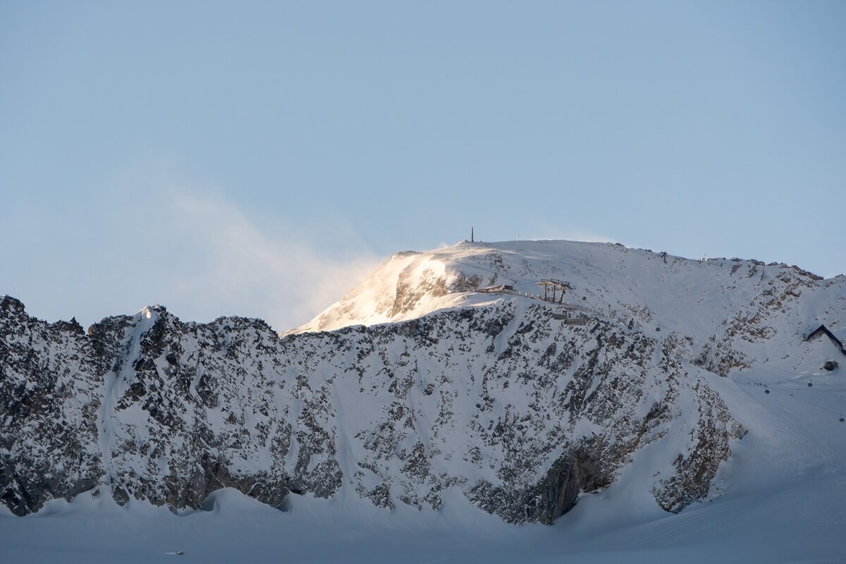 A snowy ridge with a summit station on top, lit by low sun, with wind blowing snow off the crest