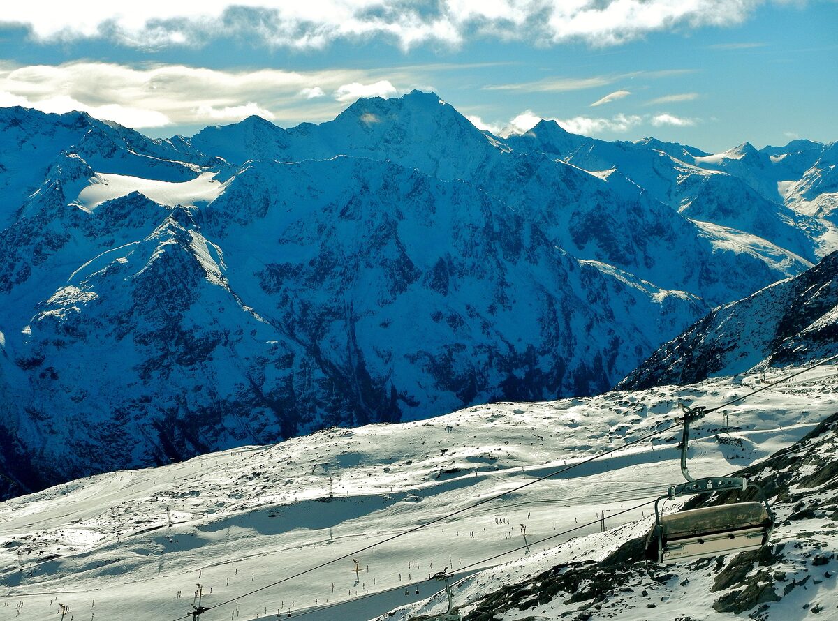 A chairlift chair above the wide snowy Tiefenbach glacier slope, with tiny skiers below and snow-covered Ötztal peaks in the sun