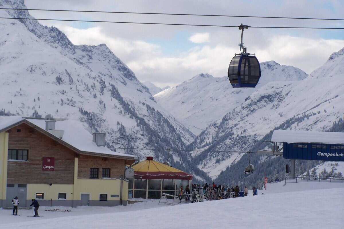 A mountain hut with a terrace at the Gampen lift station, a gondola cabin overhead and snowy peaks behind