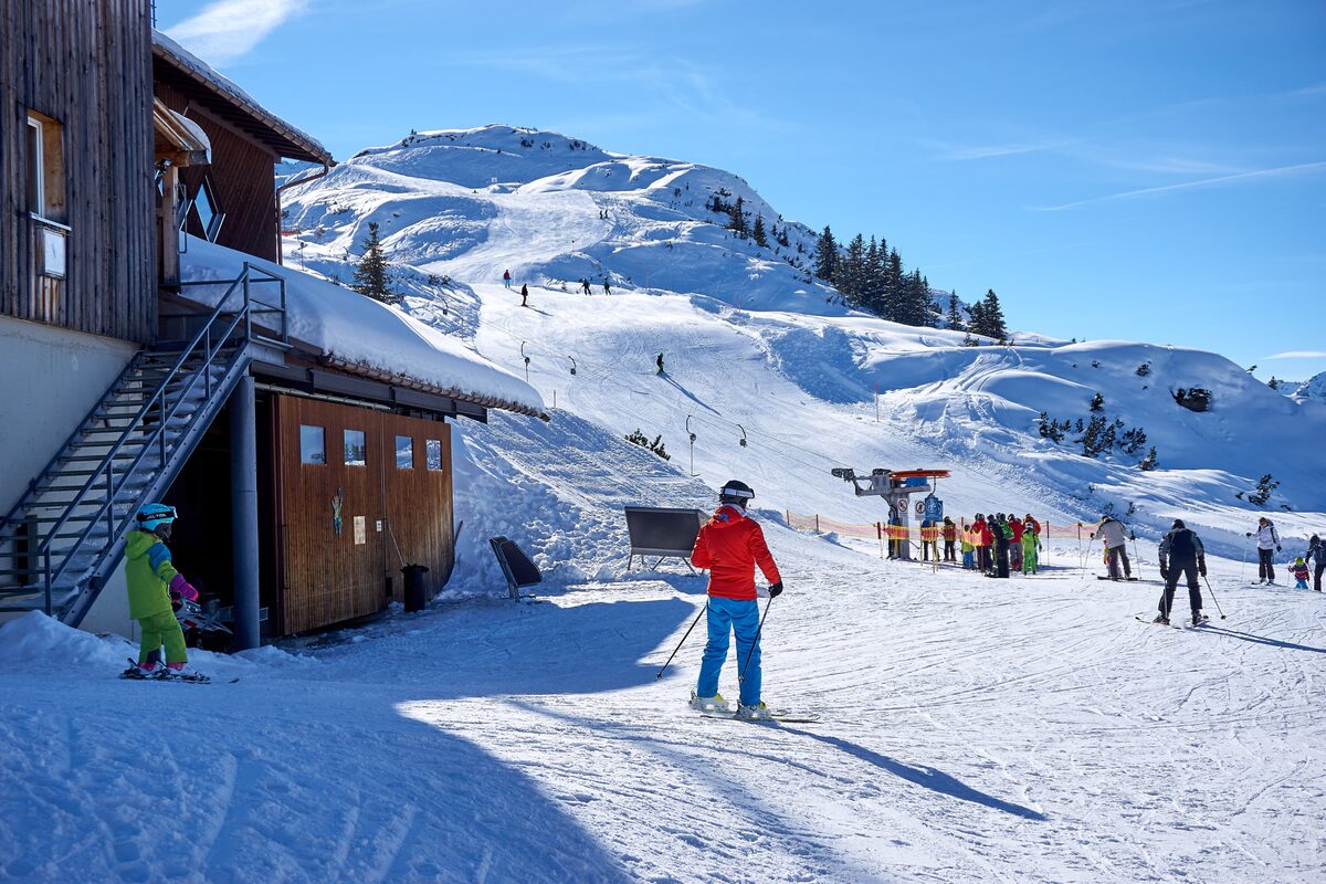 Skiers on a sunny piste at the Sonnenkopf ski area, beside a wooden mountain restaurant