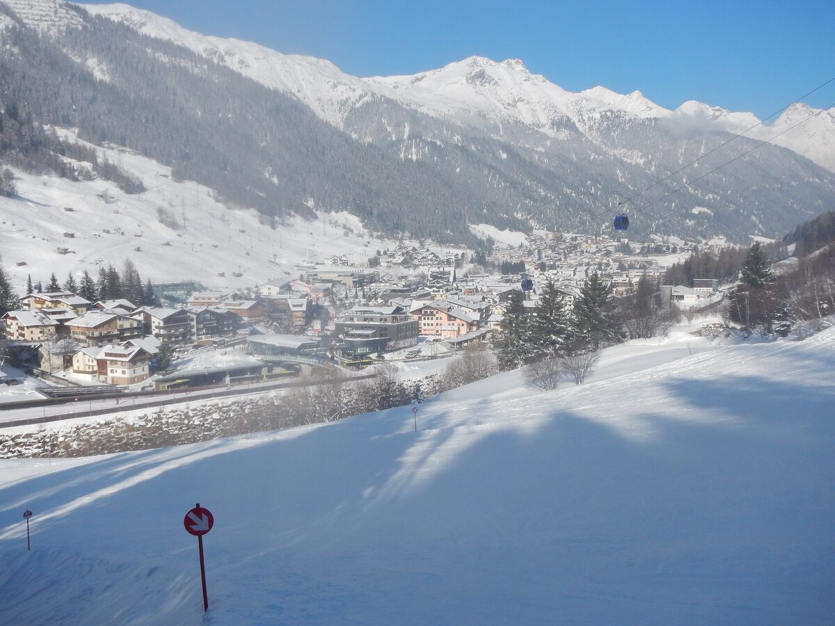 The village of St. Anton in its snowy valley seen from a sunny piste, with a gondola cabin and the peaks behind