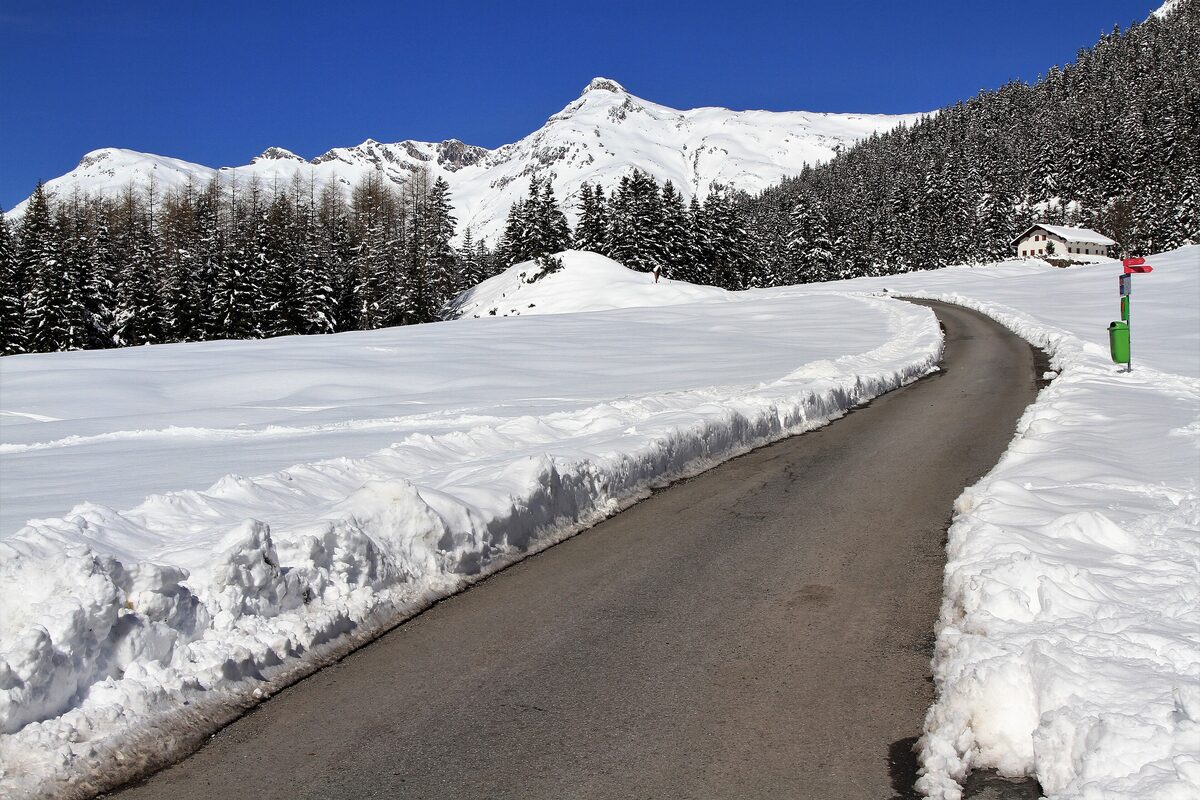 A snowbound road and deep snow banks at Zug near Lech, with a snowy peak behind the fir trees
