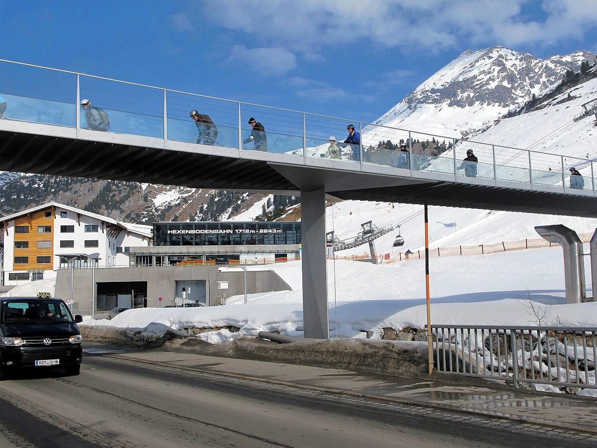 The ski bridge over the road in Zürs with skiers crossing it, the Hexenboden gondola station and snowy slopes behind