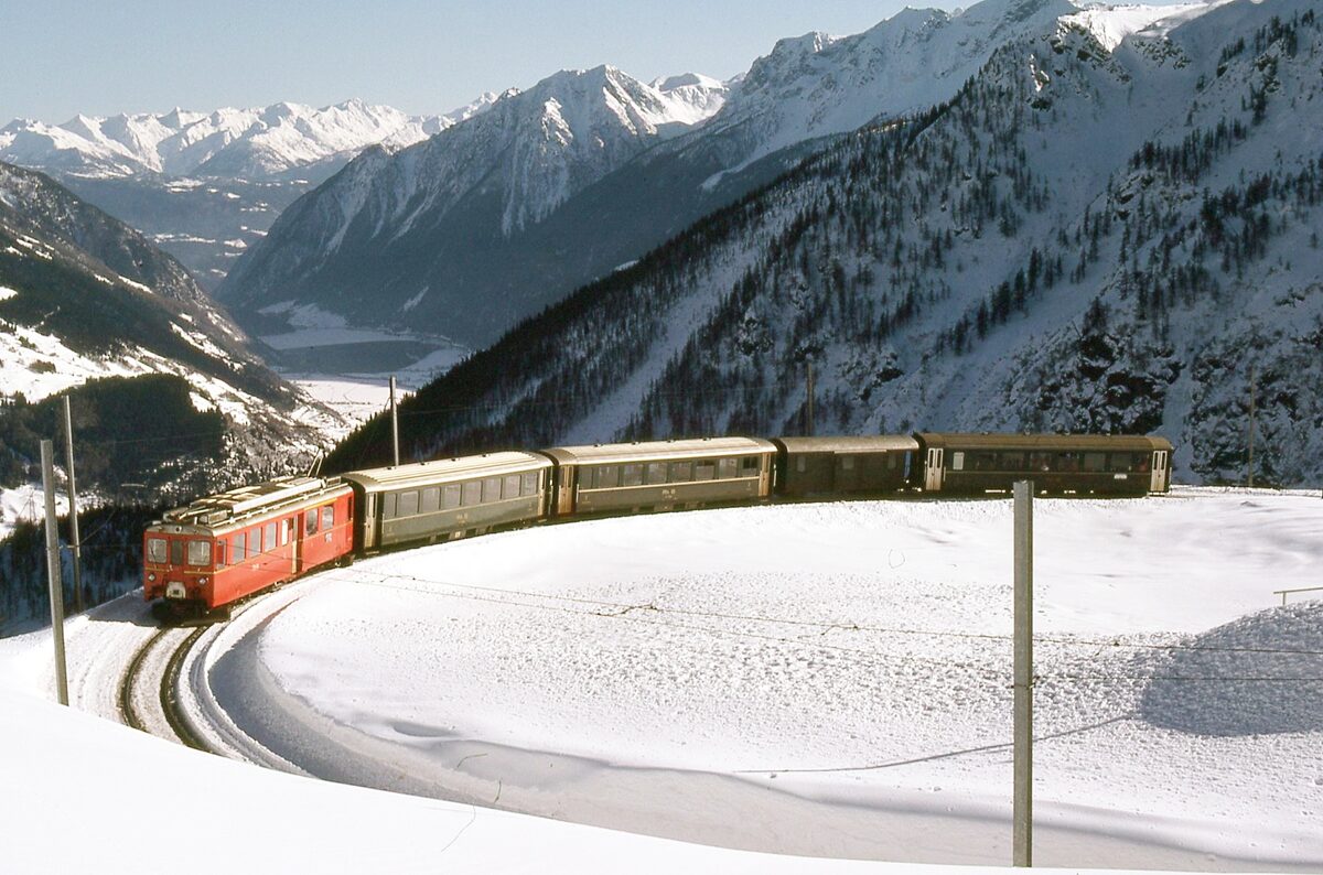 An older red Bernina line railcar with vintage carriages curves through deep snow at Alp Grüm, with the Valposchiavo far below