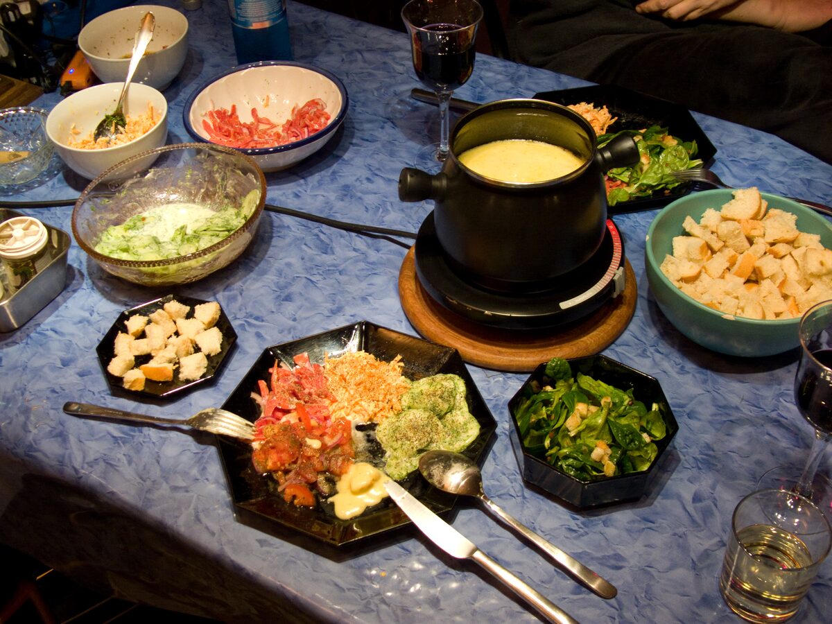 A pot of melted cheese fondue on a table, surrounded by salads, bread cubes and wine