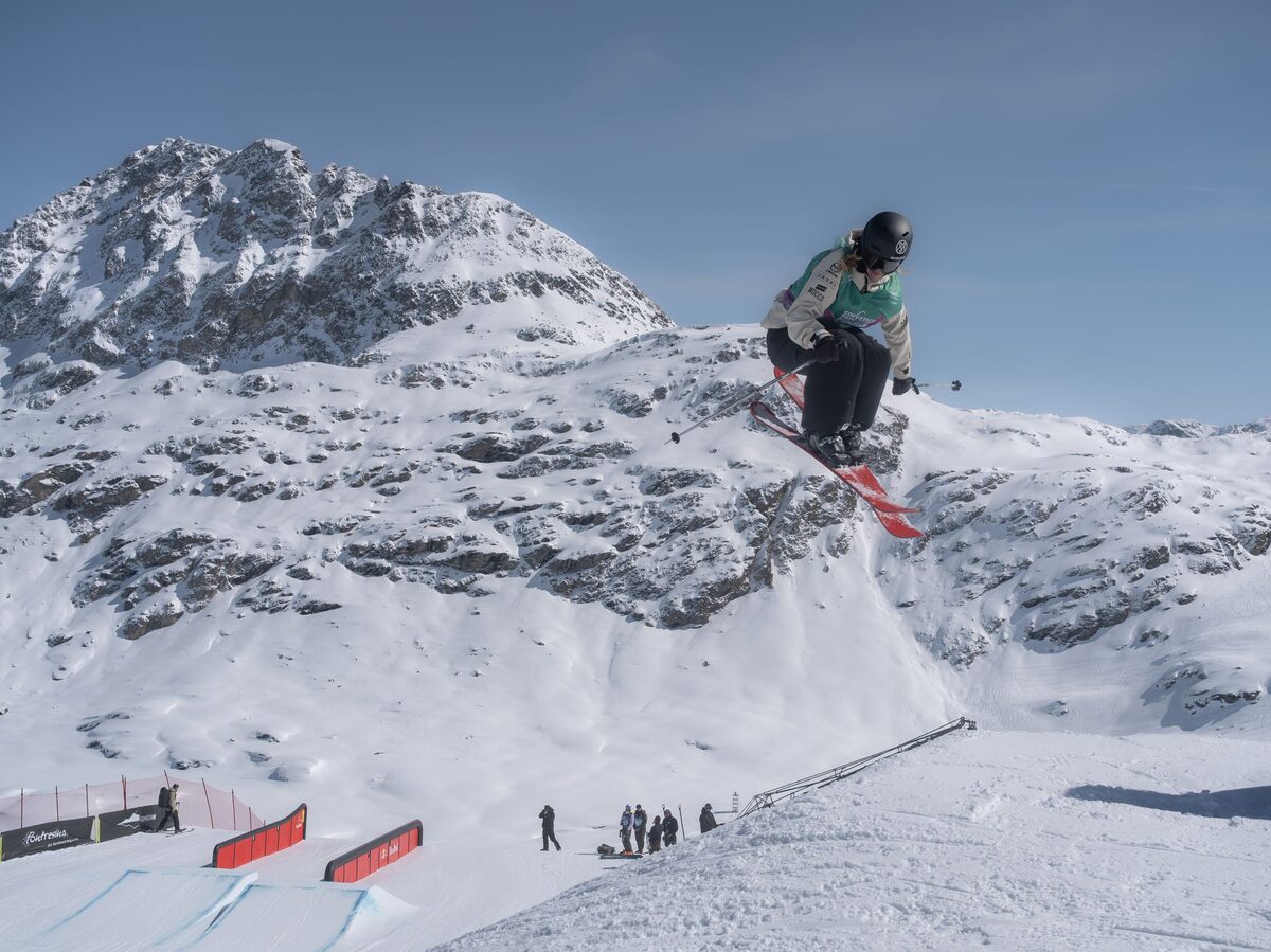 A freestyle skier takes off from a kicker in a snow park above Sils im Engadin, with rails and snowy peaks behind