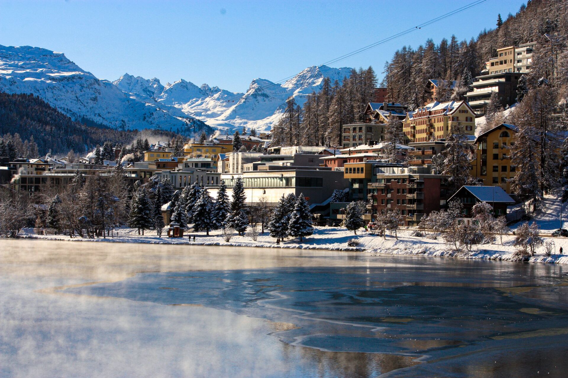 St. Moritz hotels and larch forest on the shore of the steaming, half-frozen lake, with snowy Engadin peaks behind