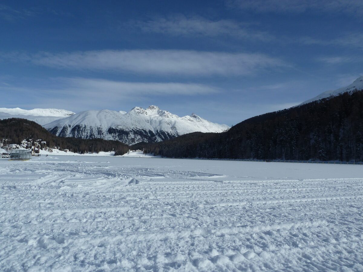 The frozen Lake St. Moritz under snow, with groomed tracks across the snow-covered ice and mountains beyond