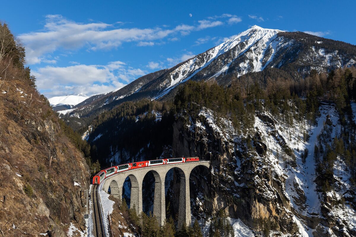A red Rhaetian Railway train on the arched Landwasser Viaduct, with snowy hillsides around