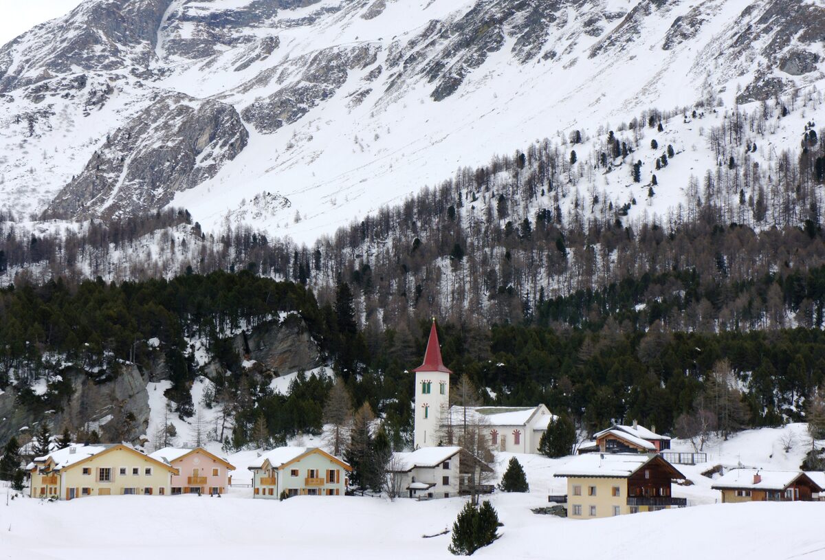 The old church and snow-covered houses of Maloja in winter, with steep wooded slopes behind