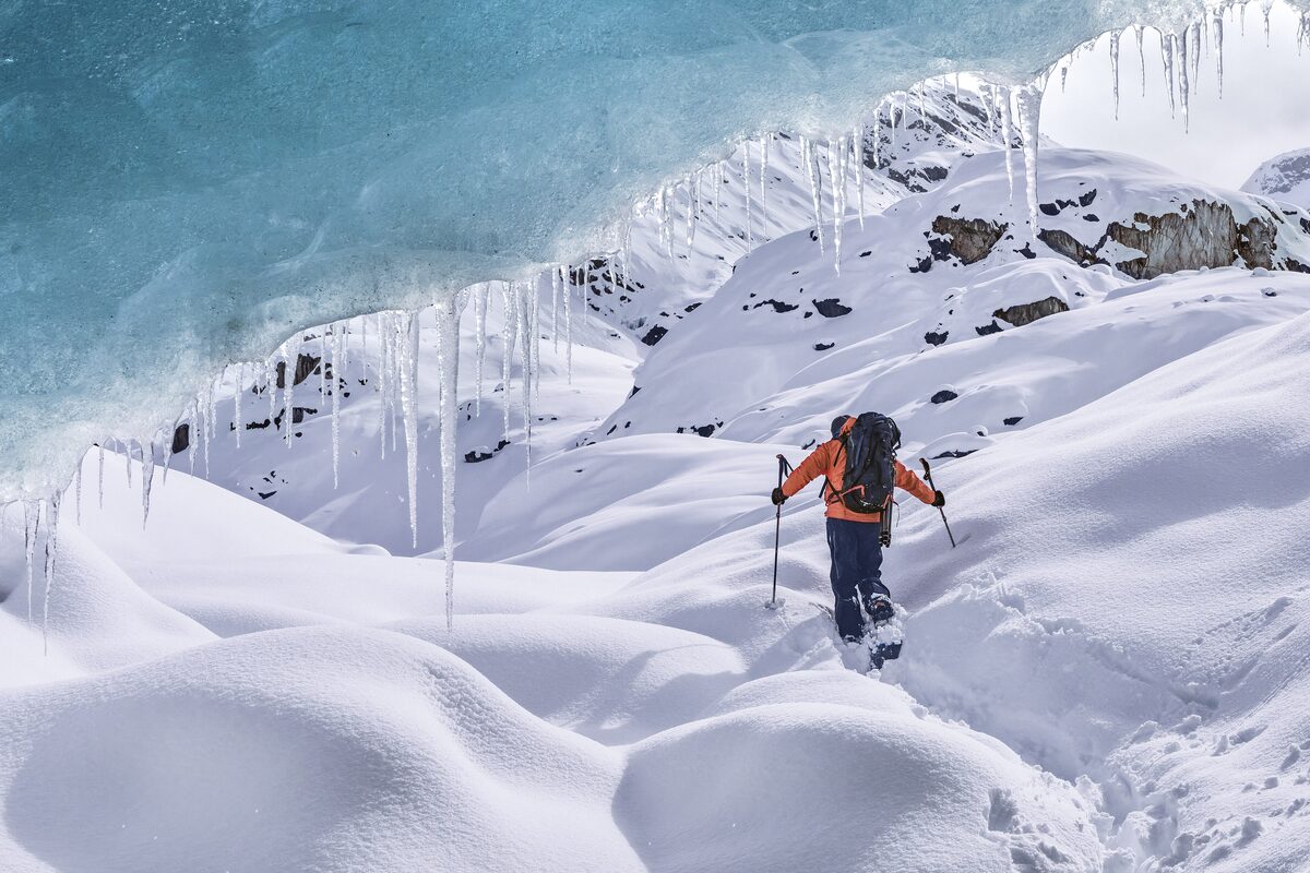A hiker with poles climbs through deep February snow beneath the blue ice of the Morteratsch Glacier