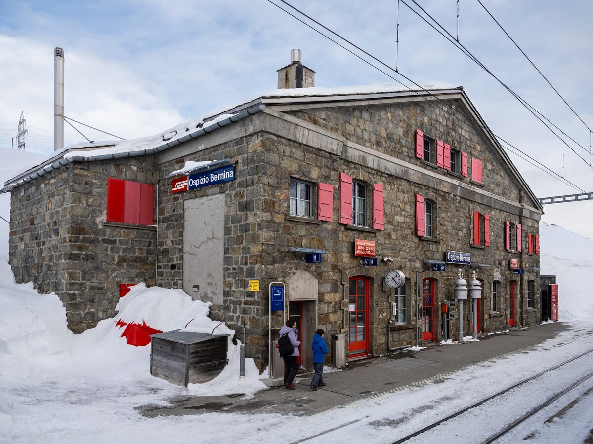 The stone station building at Ospizio Bernina in deep snow, with people on the platform