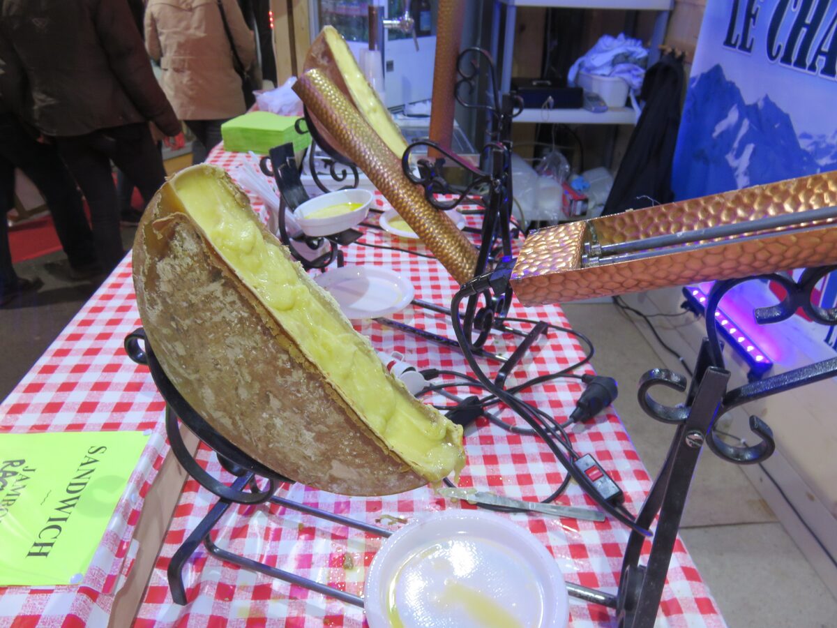 Half a wheel of raclette cheese melting under a heater at a market stall with a red checked tablecloth