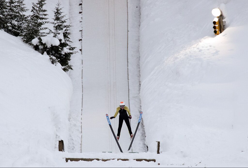 A ski jumper stands on a snowy in-run as evening light falls