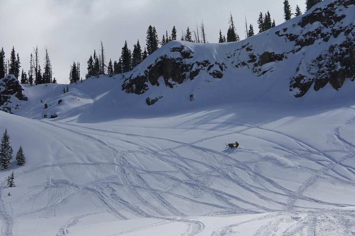 A snowmobiler's tracks crossing a wide, snowy bowl below a rocky ridge