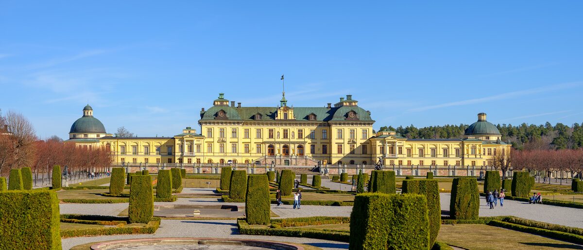 The yellow Baroque facade of Drottningholm Palace behind clipped hedges in its formal garden under a blue sky