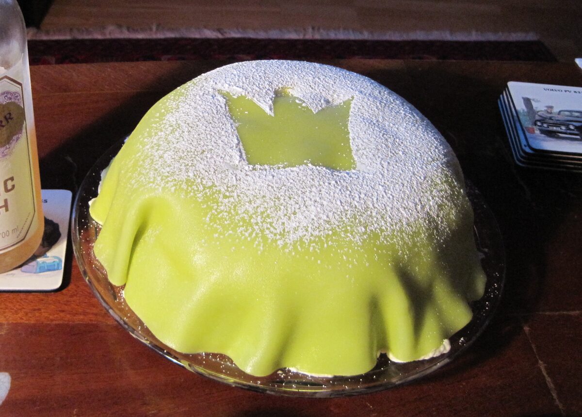 A dome-shaped princess cake covered in green marzipan with a powdered sugar crown on top