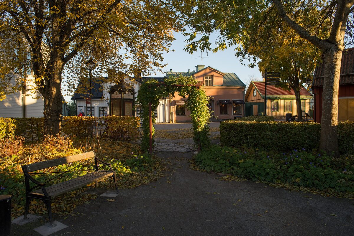 A park path with a bench under autumn trees leading to old wooden houses on Sigtuna's main street