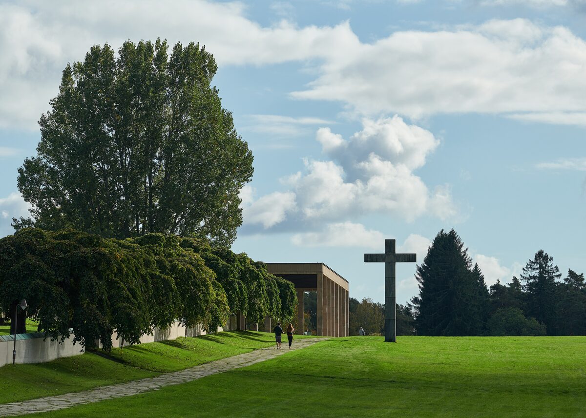 A tall granite cross on a sloping green lawn at Skogskyrkogården, with a chapel portico and trees behind