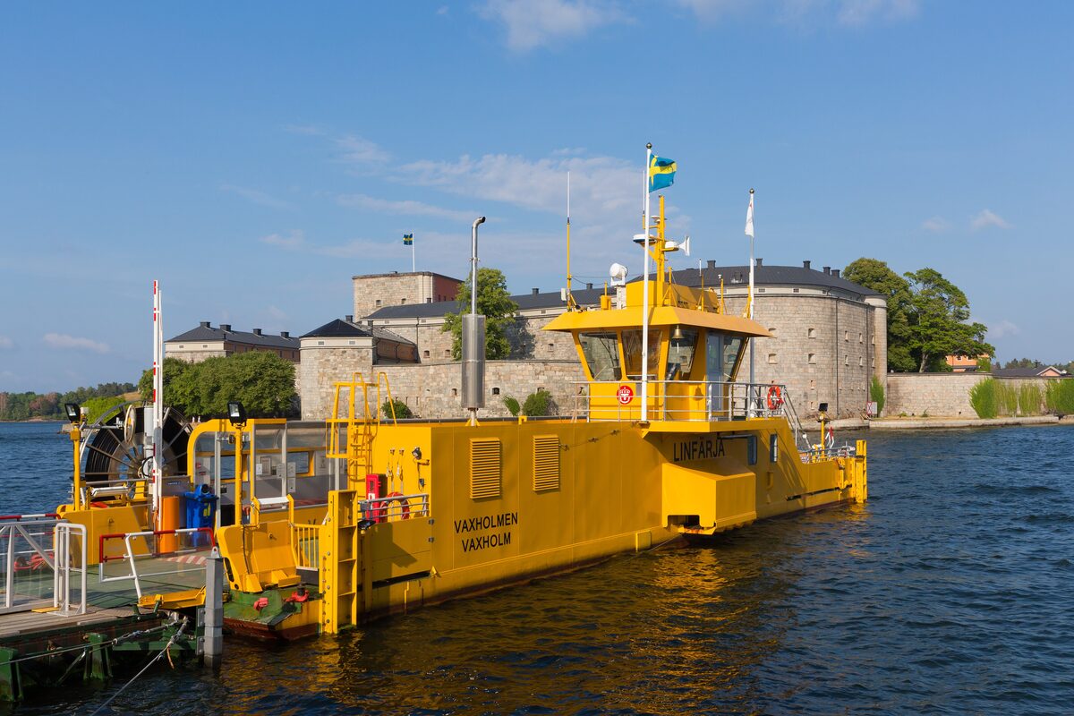 A small yellow ferry at a pier in front of the round stone Vaxholm Fortress on the water