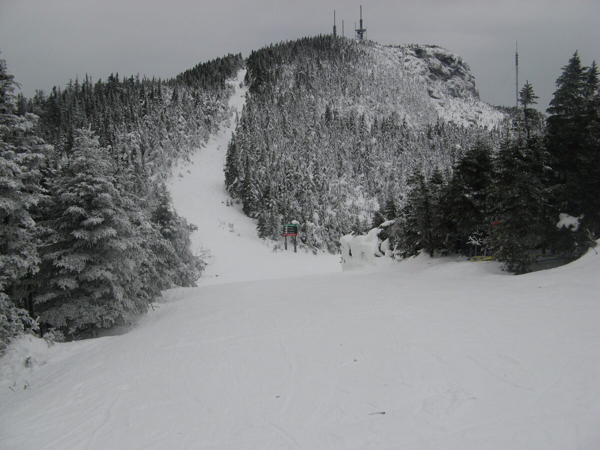 Snow-covered ski trail below a wooded Stowe summit with towers