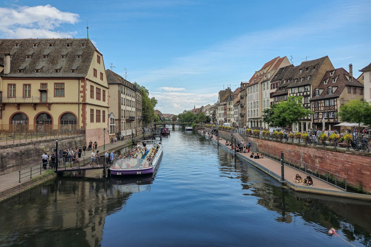 A glass-roofed sightseeing boat at its landing stage on the Ill, with old merchant houses on the left and half-timbered houses along the quay ahead