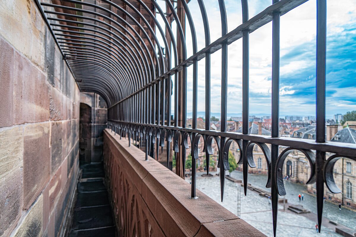 The caged stone walkway of the cathedral platform, with a wrought-iron railing and the roofs of the old town below