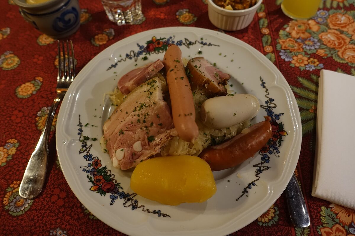 A plate of choucroute garnie with pork, sausages and boiled potatoes, with a yellow pickle on the side