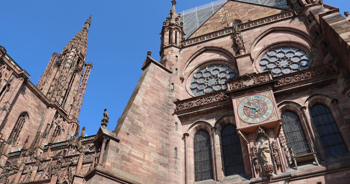 The pink sandstone west front of Strasbourg Cathedral under a blue sky, with its rose window, the clock dial on the facade and the openwork spire rising at the left