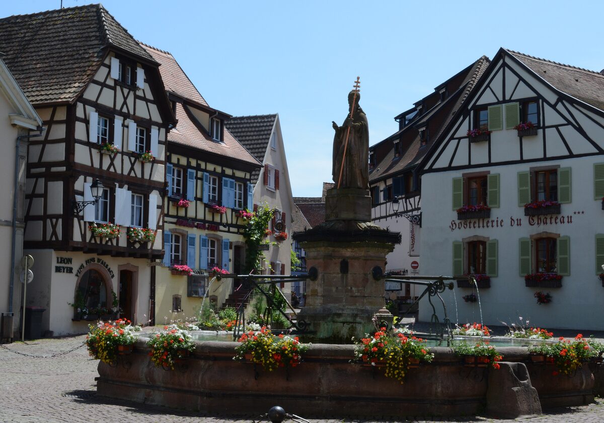 A fountain in the middle of Eguisheim's square, with half-timbered houses and red flowers in the planters