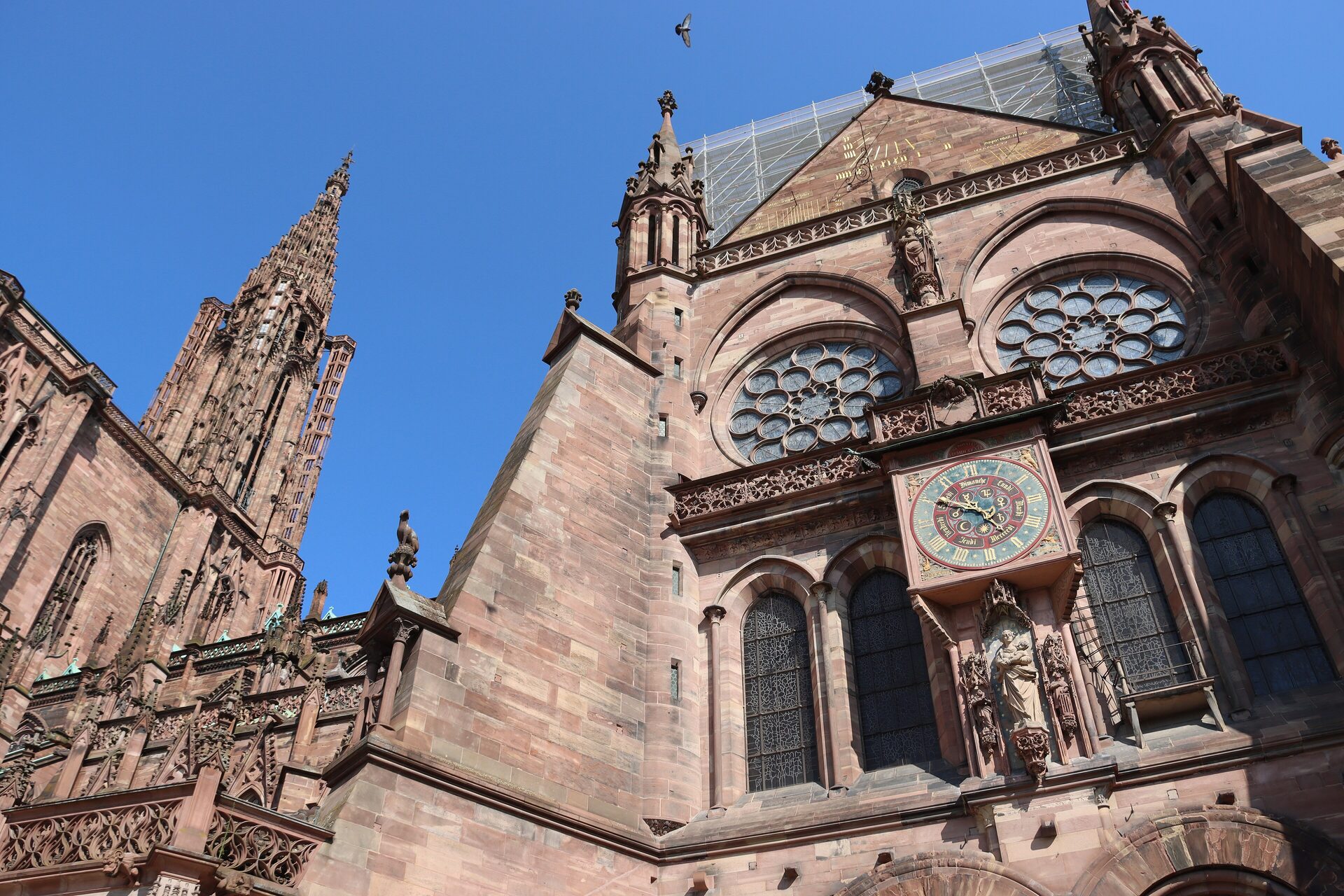 The pink sandstone west front of Strasbourg Cathedral under a blue sky, with its rose window, the clock dial on the facade and the openwork spire rising at the left