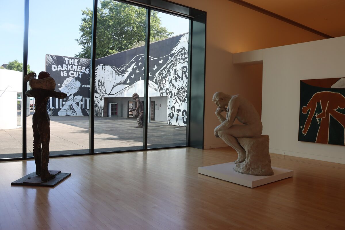 A gallery of the Museum of Modern and Contemporary Art with a bronze sculpture and a seated Rodin-style figure beside a glass wall onto the terrace
