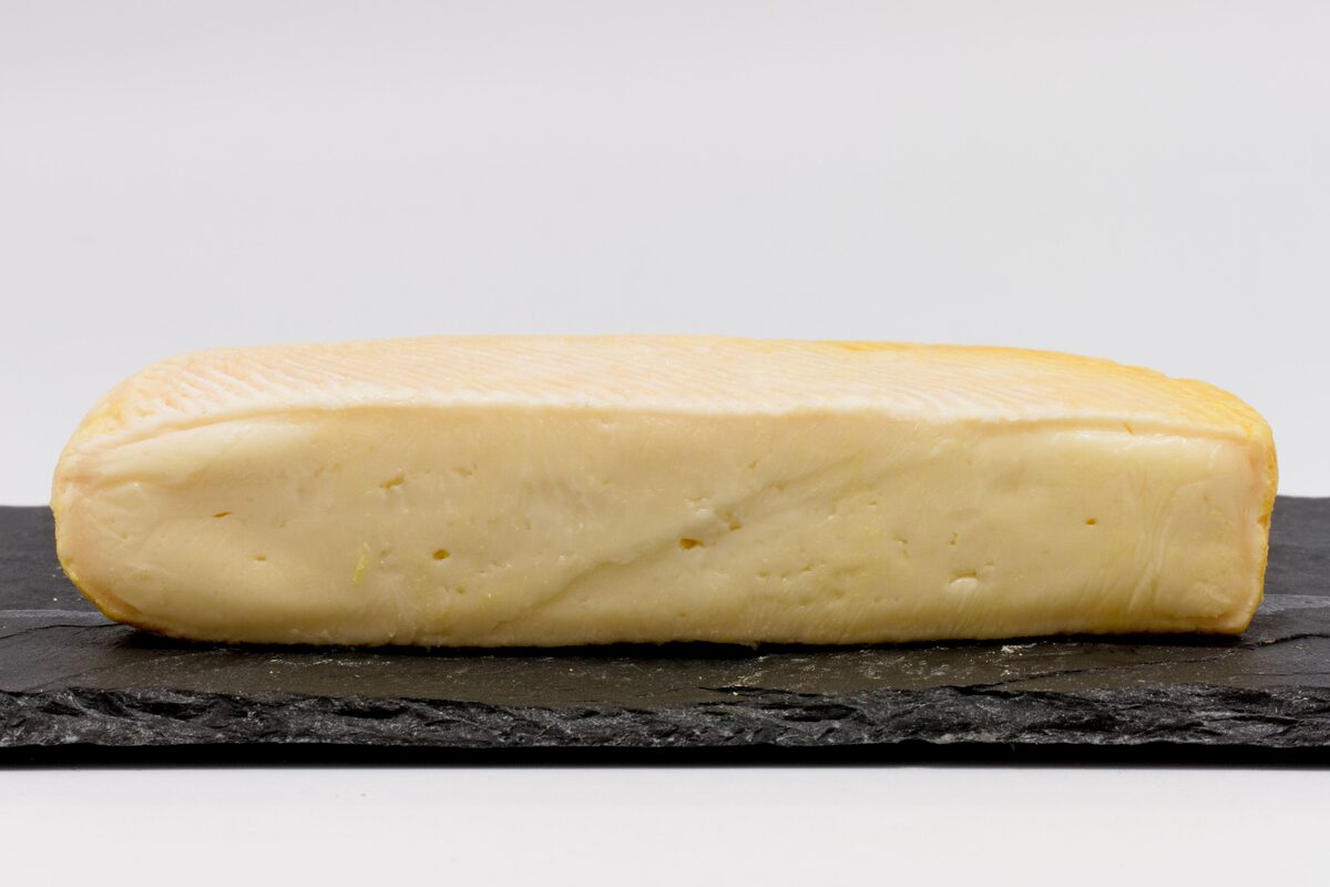 A whole Munster cheese with a pale yellow paste and thin rind, on a dark slate board