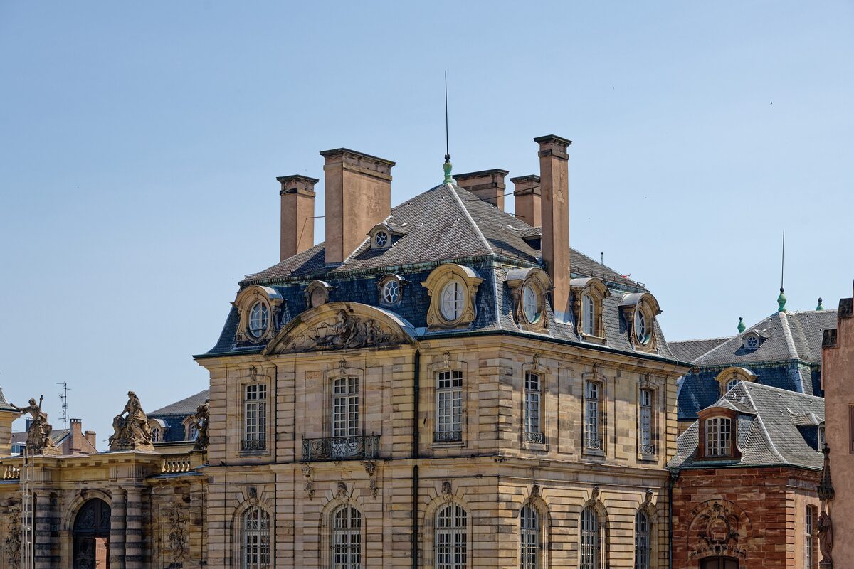 The pale stone front of the Palais Rohan under a blue sky, with a steep roof, tall chimneys and sculpted figures on the parapet