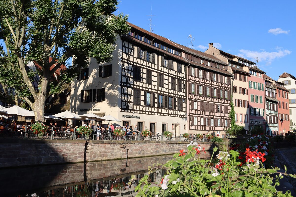 Half-timbered houses and cafe terraces along a canal in Petite France, with trees and flowers in the foreground