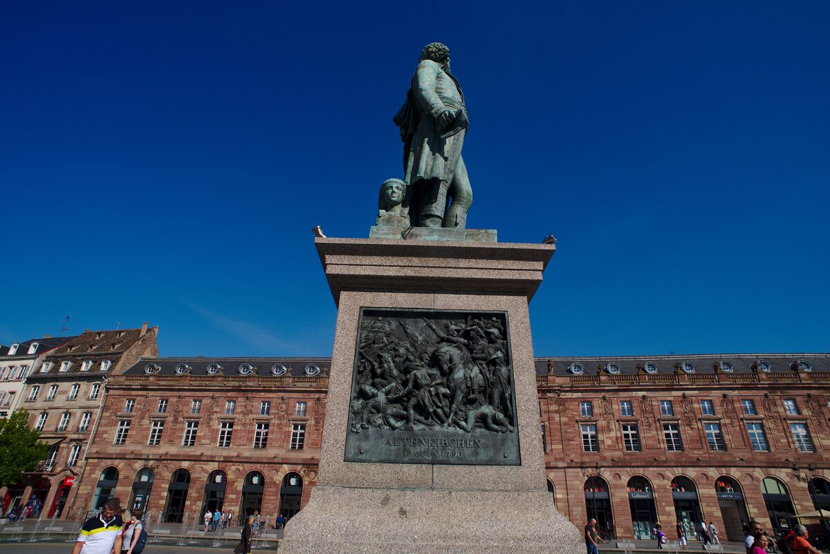 The statue of General Kléber on its stone plinth against a deep blue sky, with the long red sandstone Aubette behind it on Place Kléber