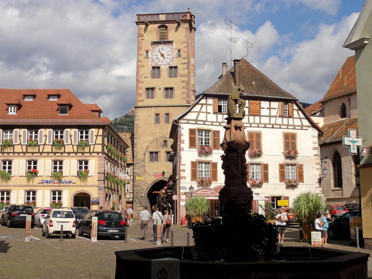 The Butchers' Tower with its clock rising over a square in Ribeauvillé, with a fountain and half-timbered houses around it
