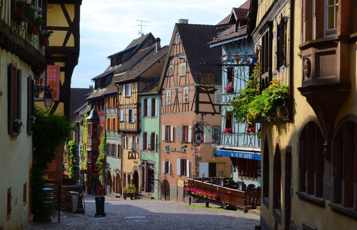 A narrow street in Riquewihr lined with half-timbered houses in pastel colors, with flower baskets on the balconies
