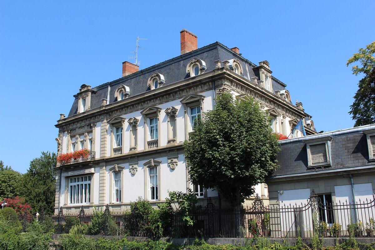 The Villa Greiner, a pale 19th-century mansion with a slate mansard roof and flower boxes, seen from its garden under a blue sky
