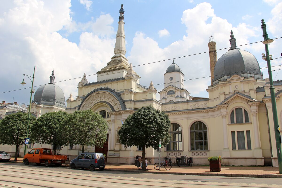 The cream-colored Anna Bath building with grey domes and a pointed central spire, behind round-clipped trees
