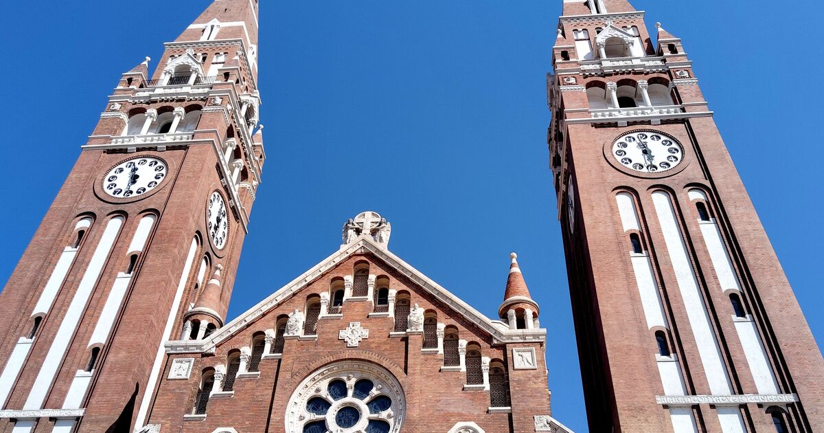 Looking up at the two brick bell towers of Szeged's Votive Church and the rose window between them, against a deep blue sky