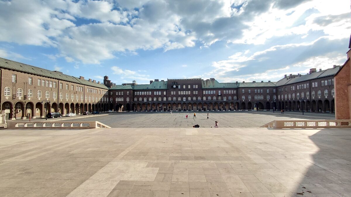 The wide paved Dóm tér, framed on three sides by long brick buildings with arcades at ground level, under a cloudy blue sky