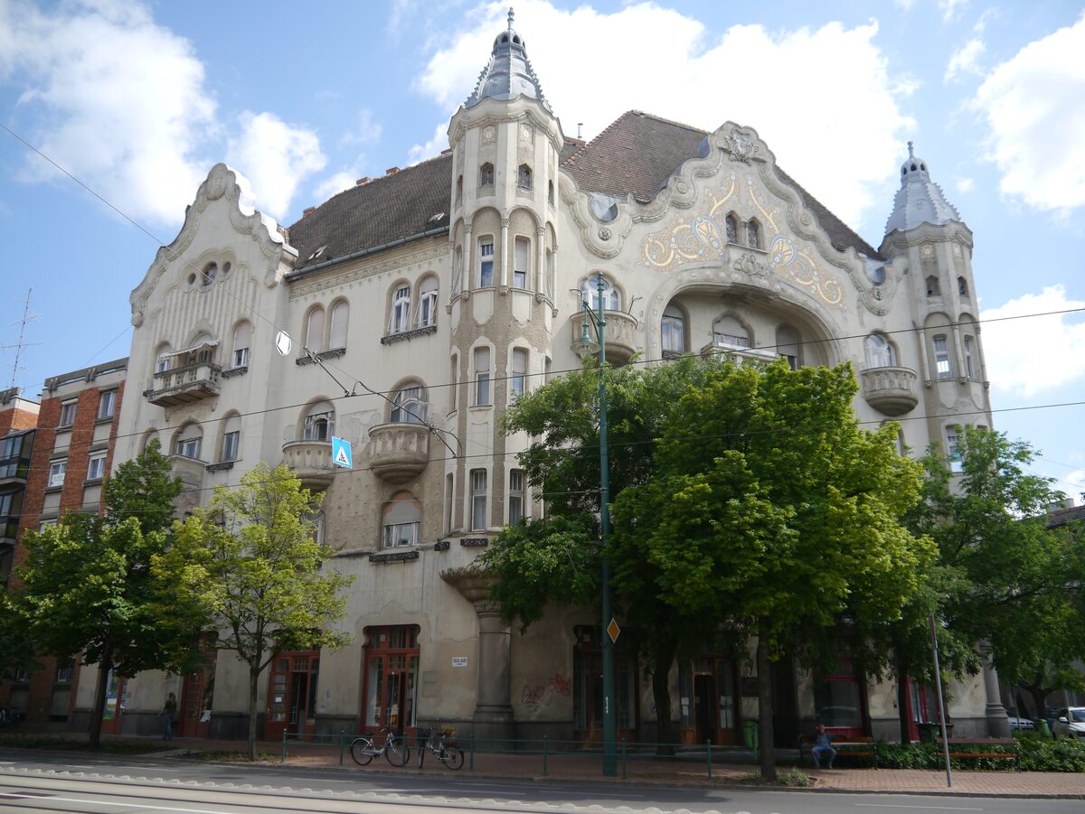The Gróf Palace, a turreted Art Nouveau apartment building with gold ornament on its facade, behind a row of green trees