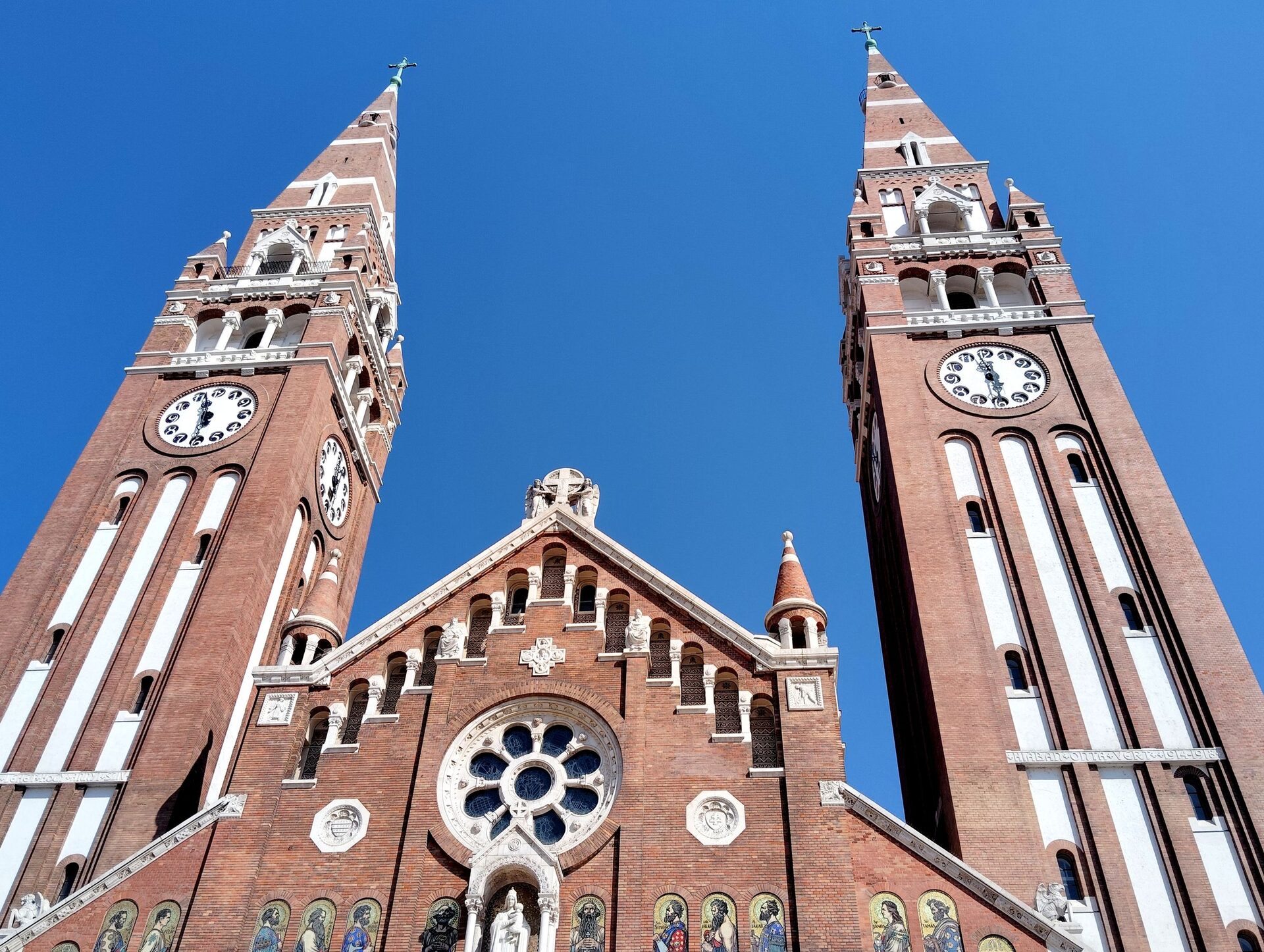 Looking up at the two brick bell towers of Szeged's Votive Church and the rose window between them, against a deep blue sky
