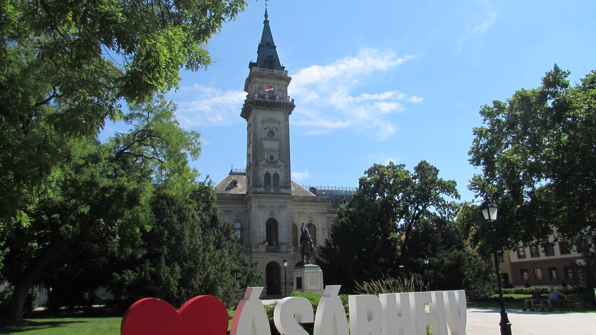 Hódmezővásárhely Town Hall's tall clock tower rising behind trees and an equestrian statue on Kossuth Square
